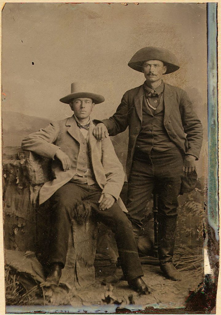 Vintage sepia-toned portrait photograph showing two men in historical clothing and cowboy hats, one seated and one standing, posed against a rustic outdoor backdrop with a mottled paper border.