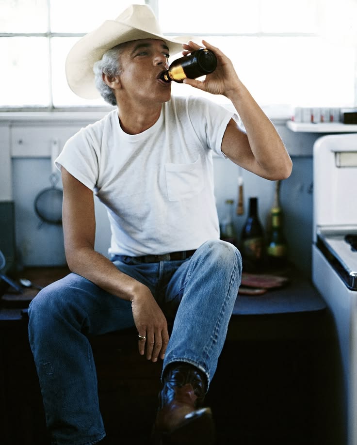 Man in a cowboy hat drinking from a bottle in a kitchen.