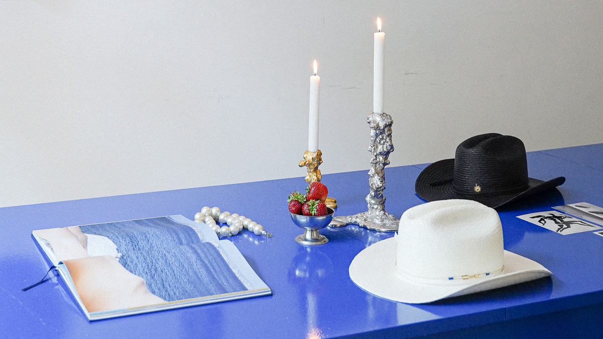 A styled tabletop arrangement on a bright blue surface against a light gray wall, featuring a white cowboy hat, a black cowboy hat, a white candle in a silver holder, a white candle in a gold holder with strawberries, a magazine, a strand of white beads, and a small decorative bowl.