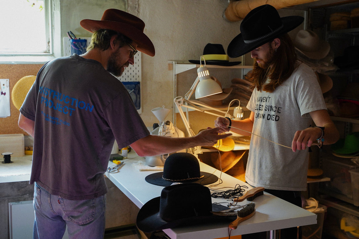 Two men in a hat-making workshop. One man on the left wears a brown cowboy hat and a gray t-shirt with blue text on the back, while the other man on the right wears a black cowboy hat and a white graphic t-shirt.
