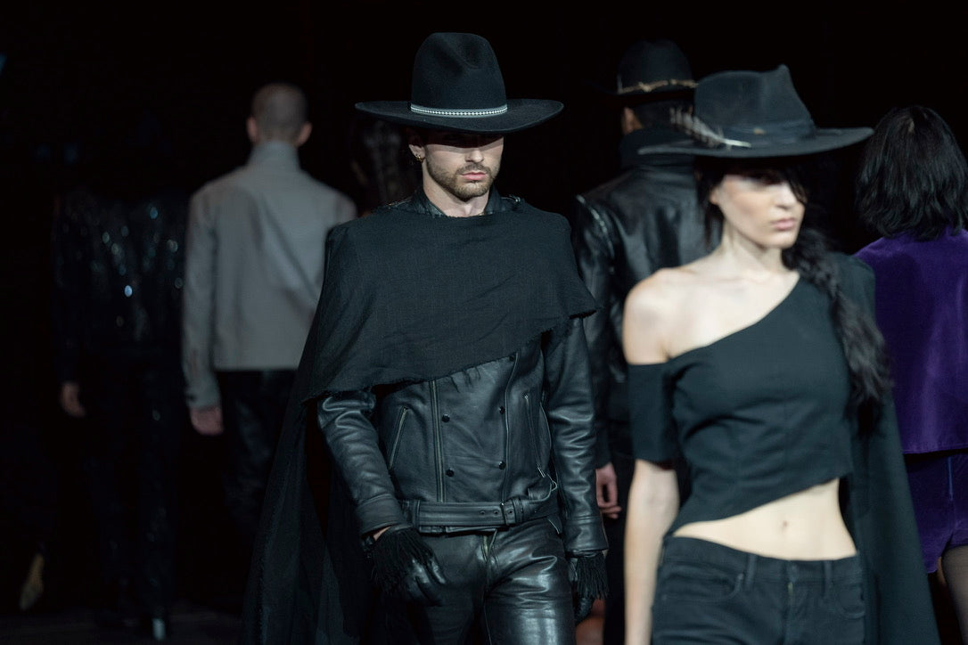 A runway fashion photo showing a model in a black leather outfit with a wide-brim black hat and cape, alongside another model in a black off-the-shoulder top and matching hat, on a dark runway.