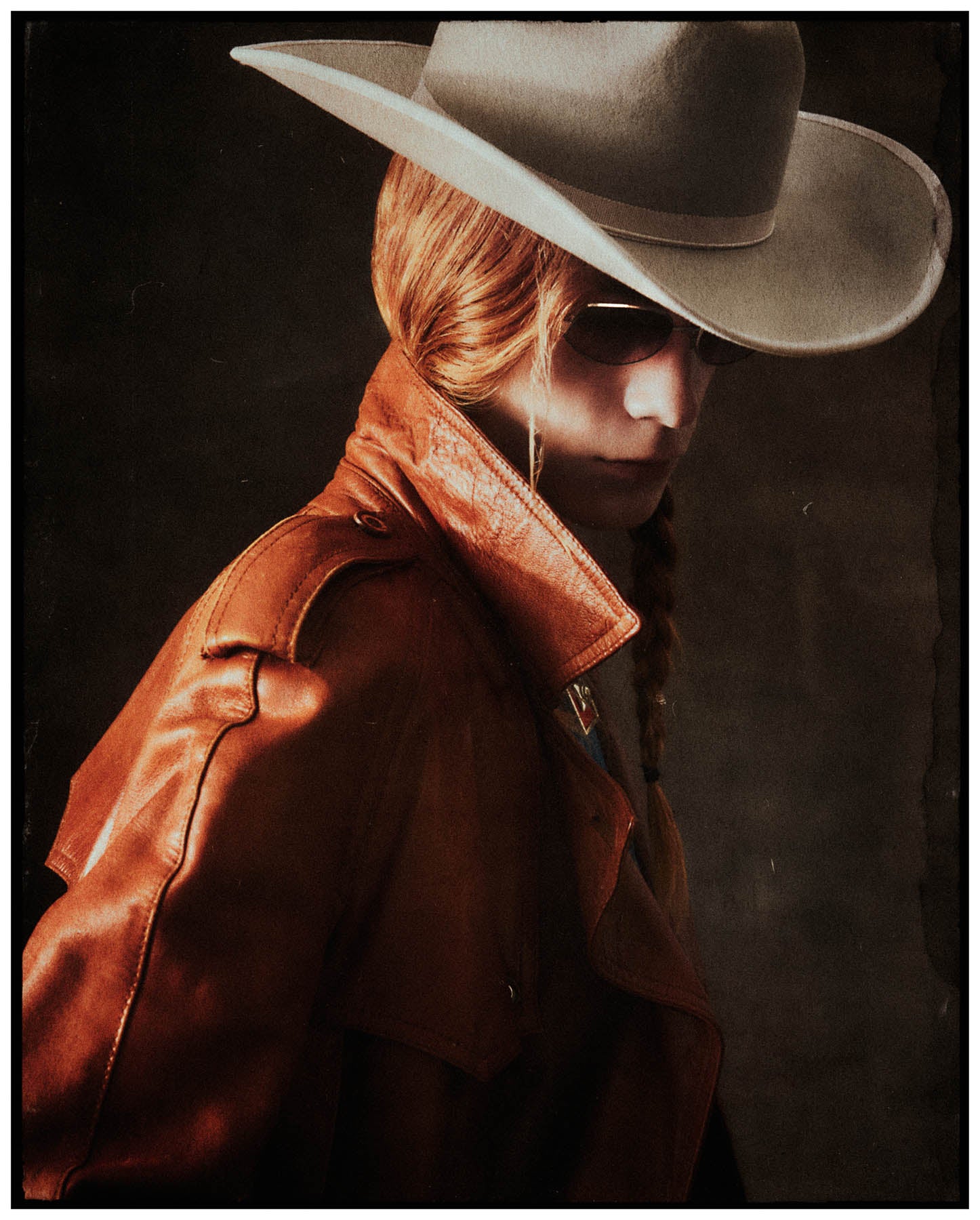 Person wearing a brown leather jacket and wide-brimmed light beige cowboy hat against a dark background
