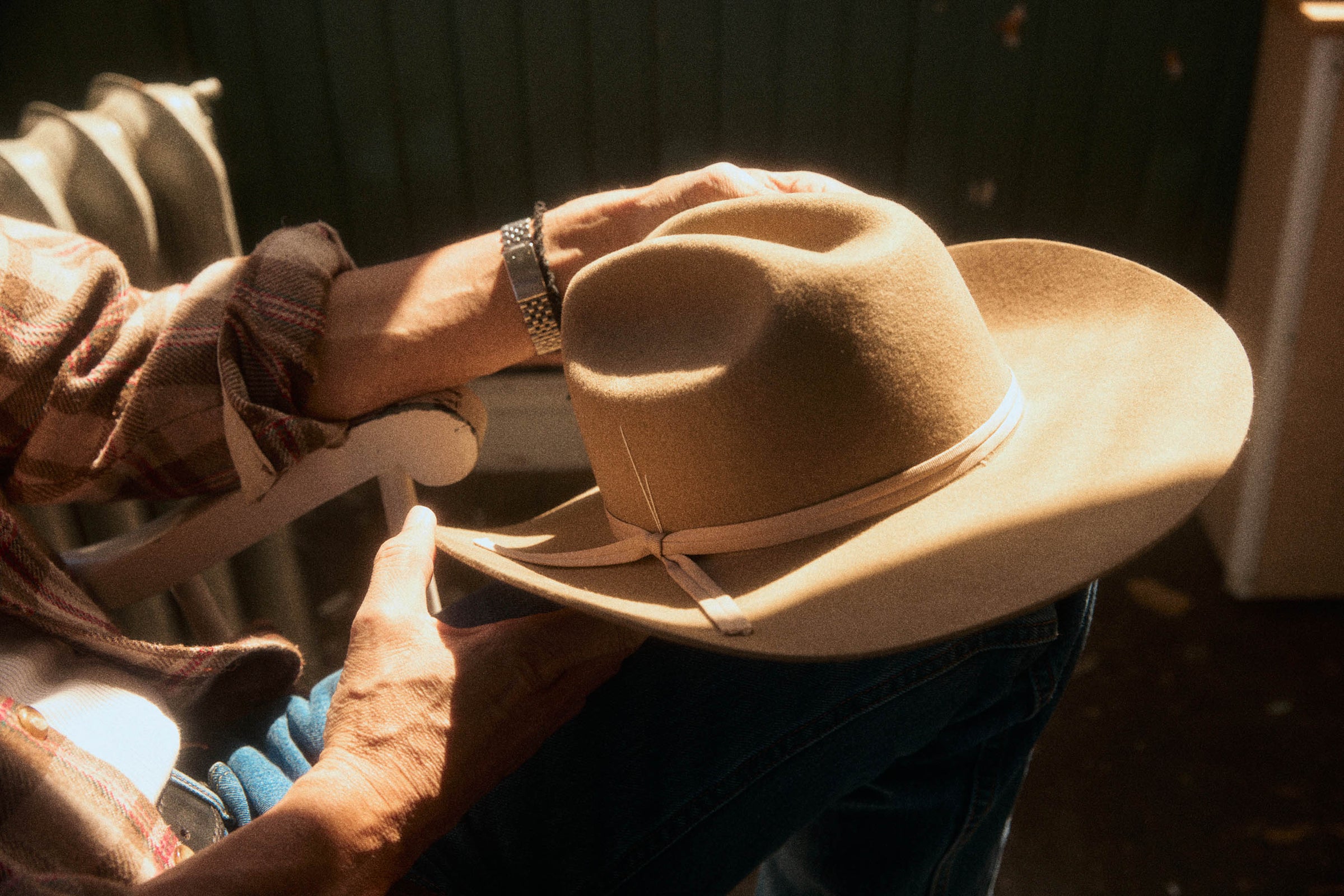 A person wearing a brown plaid shirt and blue jeans holds a light brown felt cowboy hat with a matching ribbon band in warm sunlight.