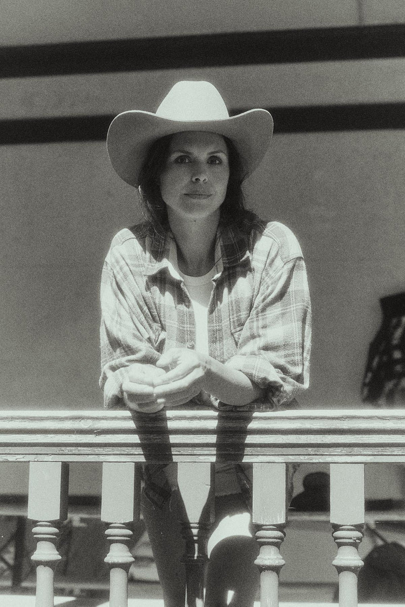 A black and white photograph of a woman wearing a cowboy hat, a plaid button-up shirt, and tall boots, leaning against a wooden railing.