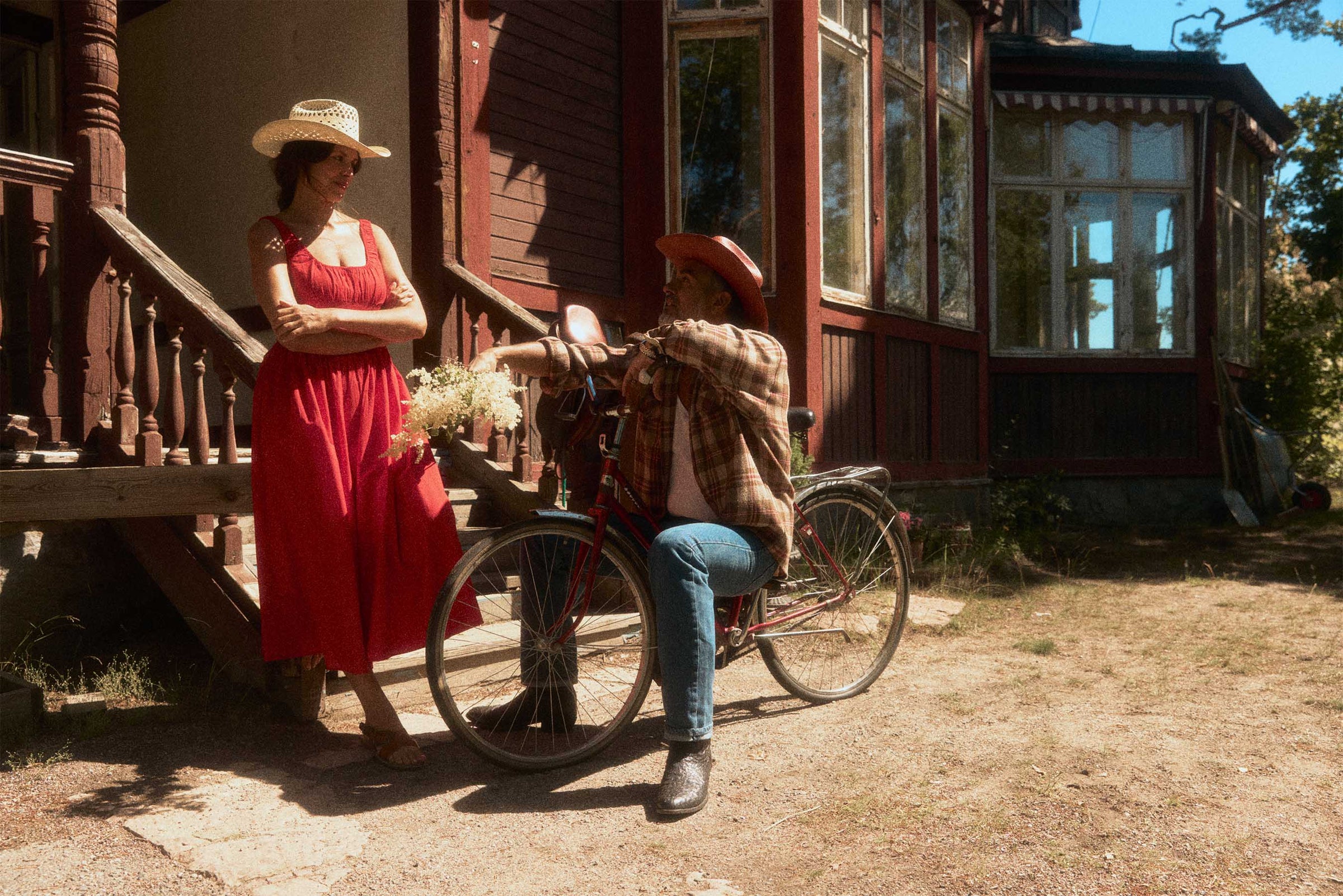 A woman in a red dress and straw cowboy hat stands on a porch next to a man sitting on a bicycle, wearing a reddish straw cowboy hat, brown plaid shirt, and blue jeans in front of a rustic red wooden house.