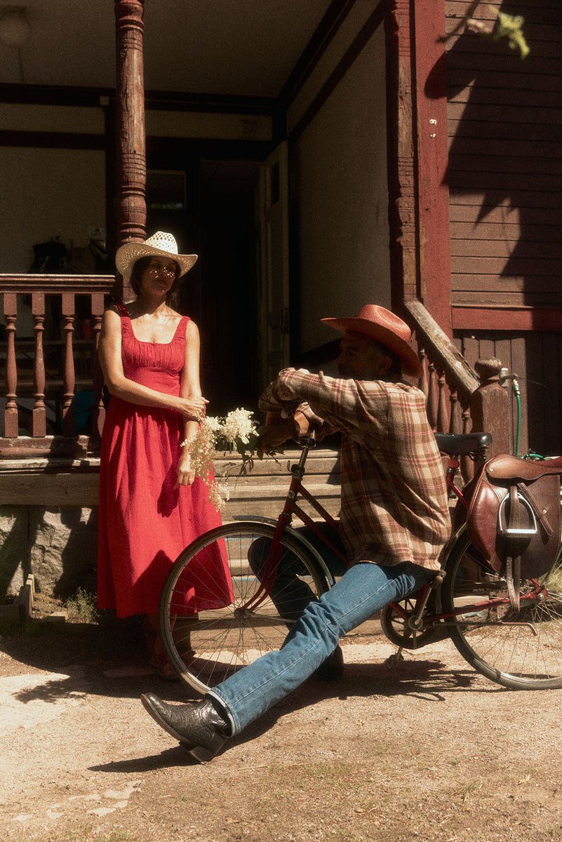 A man in a straw cowboy hat and plaid shirt leans against a bicycle on a porch while a woman in a red dress and white straw cowboy hat stands nearby in a rustic outdoor setting.