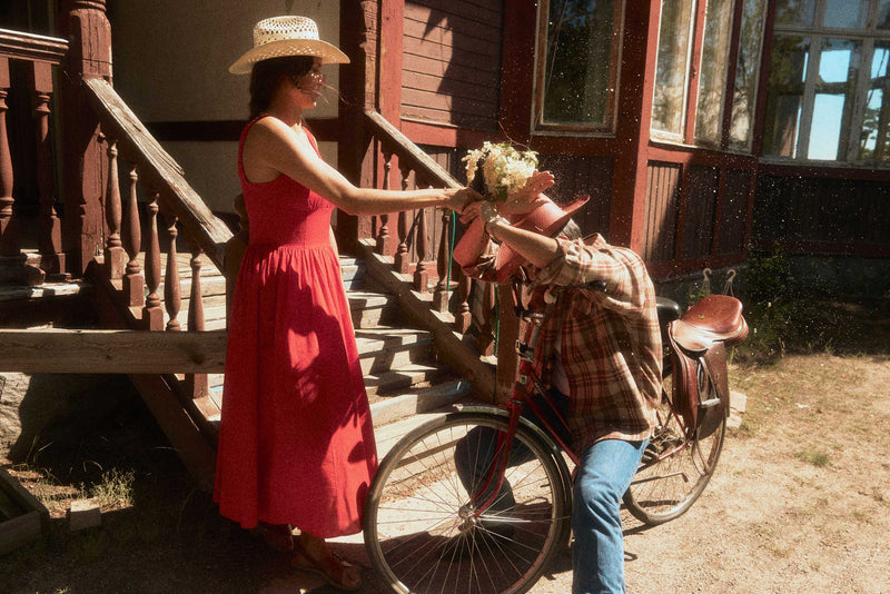 A woman in a red dress and straw cowboy hat stands on a wooden porch punching flowers on a man wearing a reddish straw cowboy hat and riding a bicycle in front of a rustic red house.
