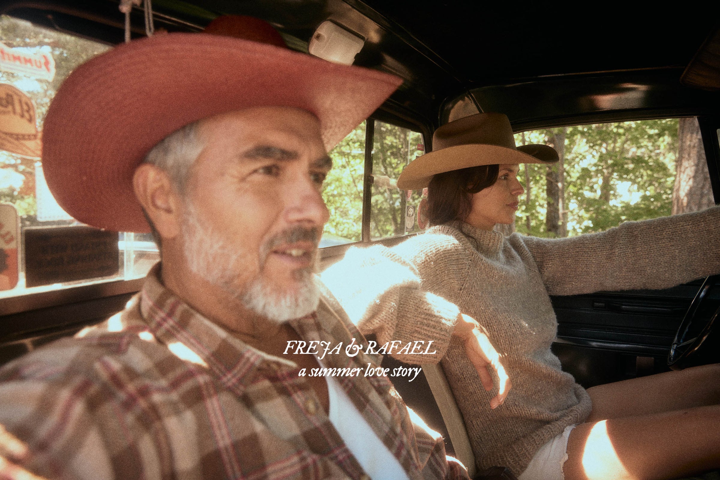 A man and a woman wearing cowboy hats sit inside a vintage car, with warm sunlight streaming through the windows. The man in the foreground wears a red cowboy hat and a brown plaid shirt, while the woman behind him wears a beige hat and a beige knit sweater.