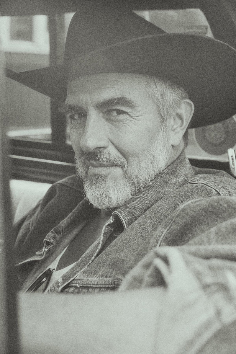 A black and white portrait of an older man with a beard wearing a wide-brimmed cowboy hat and a denim jacket, seated in a car.
