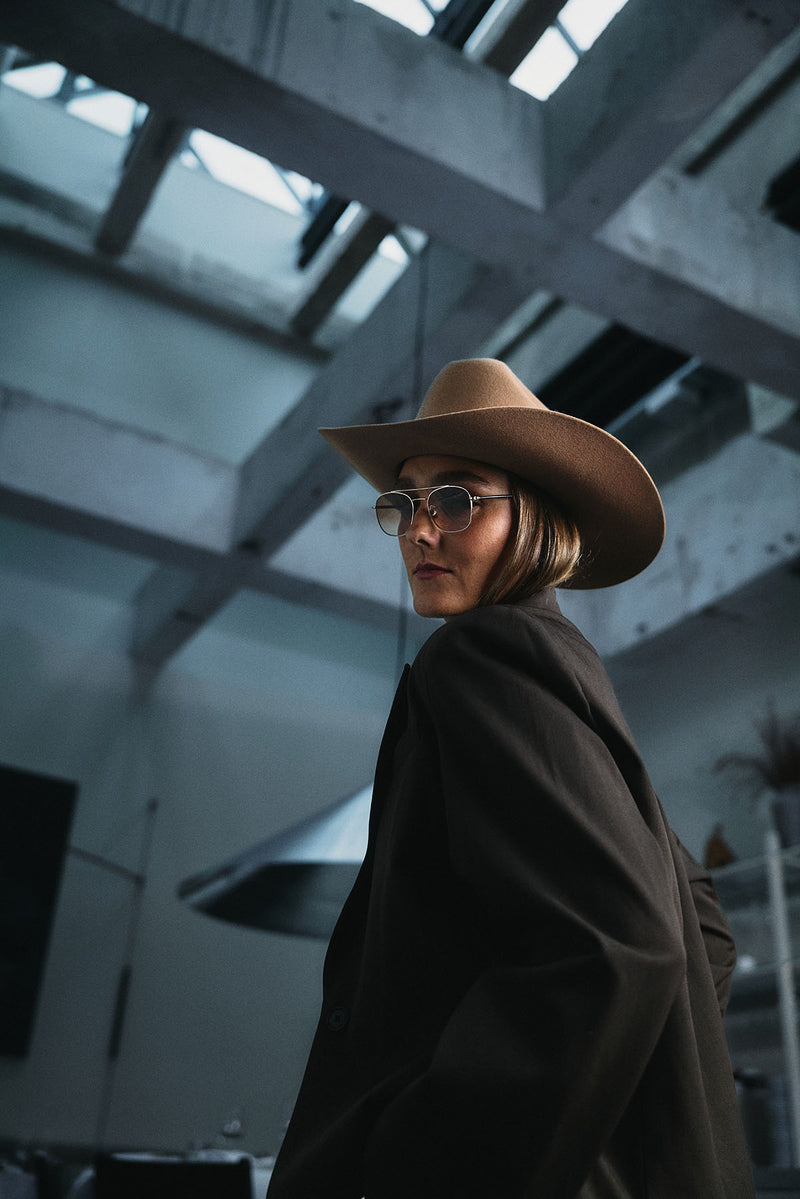 A woman wearing a wide-brim tan cowboy hat, round sunglasses, and a dark oversized coat, photographed in a high-ceilinged industrial interior with concrete beams.