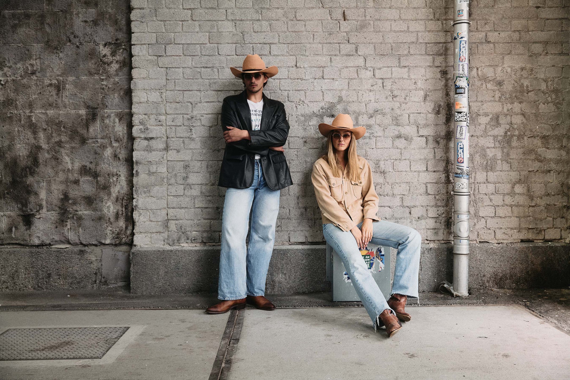 Two people in a western-inspired outfit, including cowboy hats, sunglasses, blazers, and light blue jeans, posing against a gray brick wall.