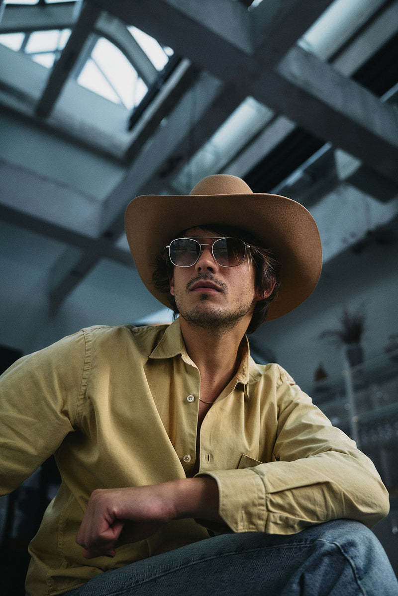 A man wearing a tan cowboy hat, sunglasses, and a yellow button-down shirt sits in an industrial loft space with high ceilings and steel beams.