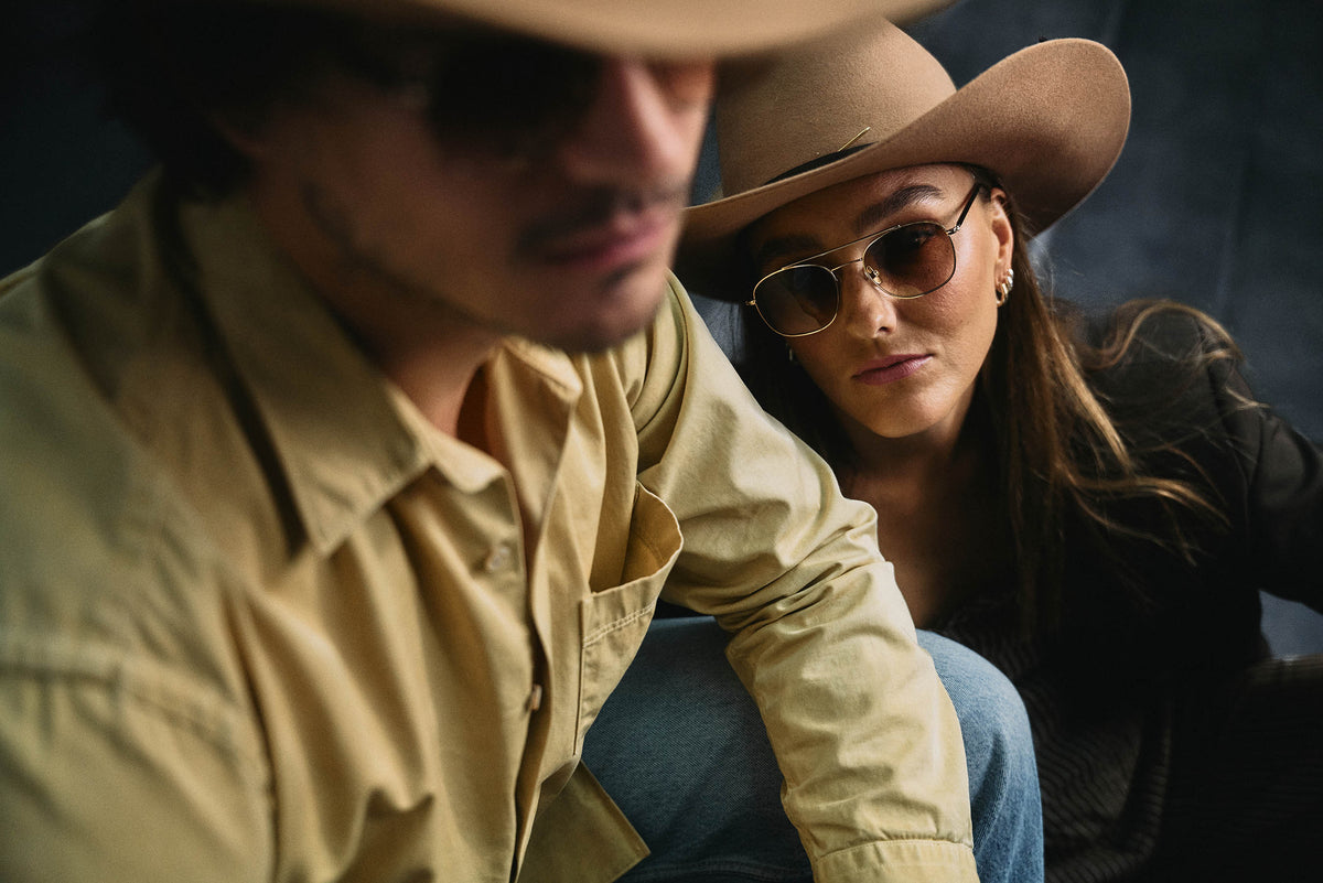 A close-up lifestyle shot of a couple wearing western-style eyewear. The woman on the right wears round gold-rimmed sunglasses with a beige cowboy hat, while the man on the left is in sharp focus wearing a tan button-down shirt and a matching cowboy hat.