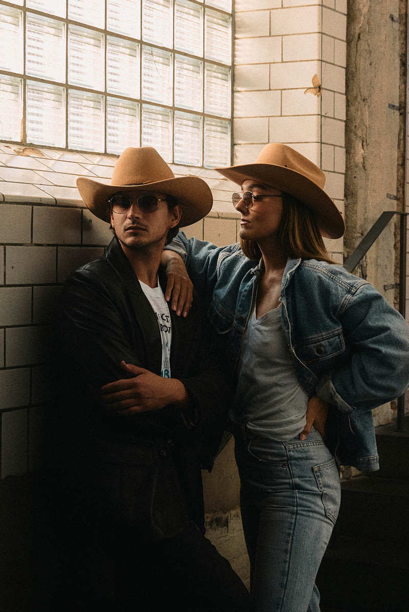 A man and a woman posing together in a rustic tiled room. The woman is wearing a tan cowboy hat, a blue denim jacket, and light blue jeans, with her arm around the man's shoulder, who's wearing a tan cowboy hat and black leather jacket.