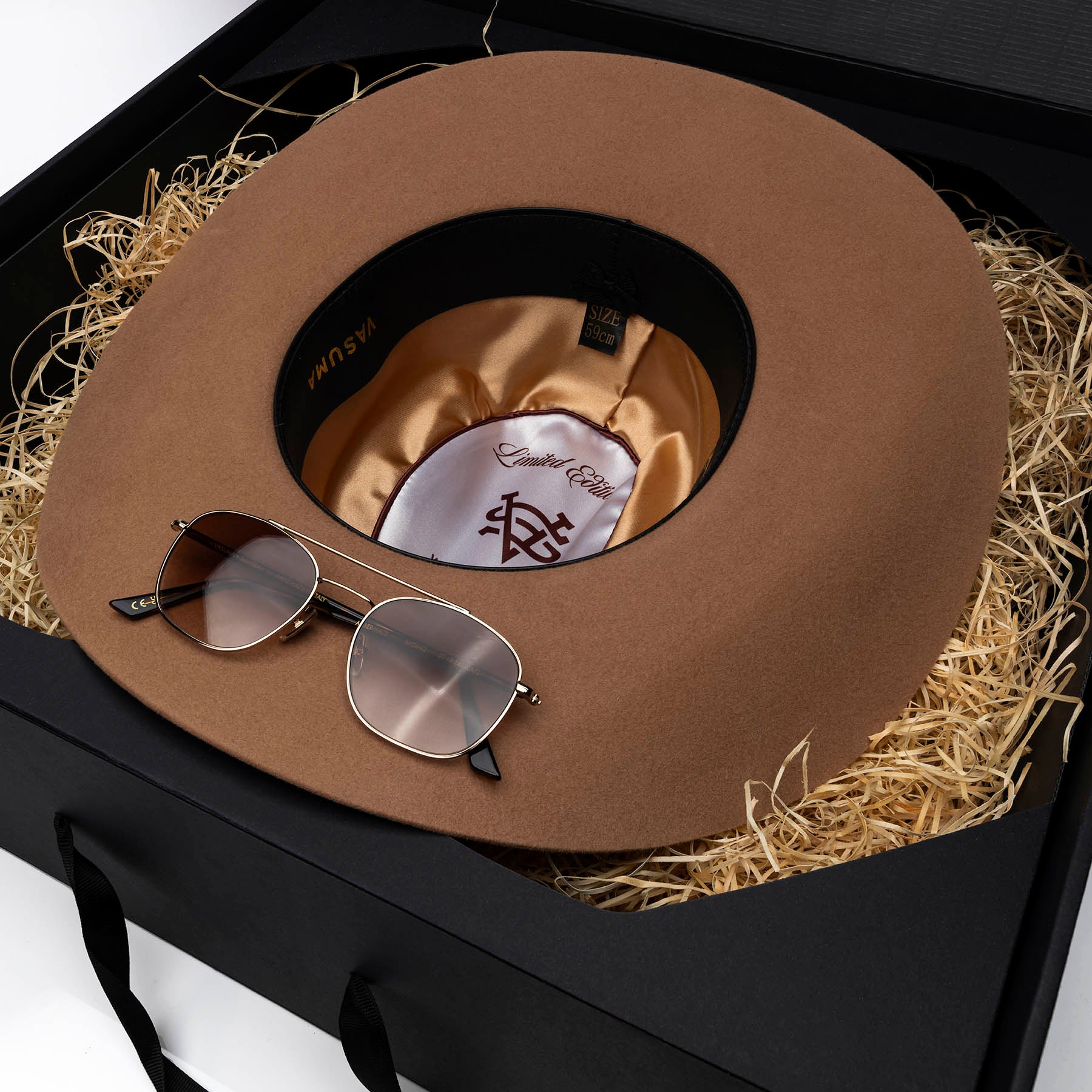 A brown wide-brim cowboy hat displayed in a black gift box with wood straw filler, alongside a pair of aviator sunglasses. The hat features a black leather sweatband with gold lettering and a satin inner lining.