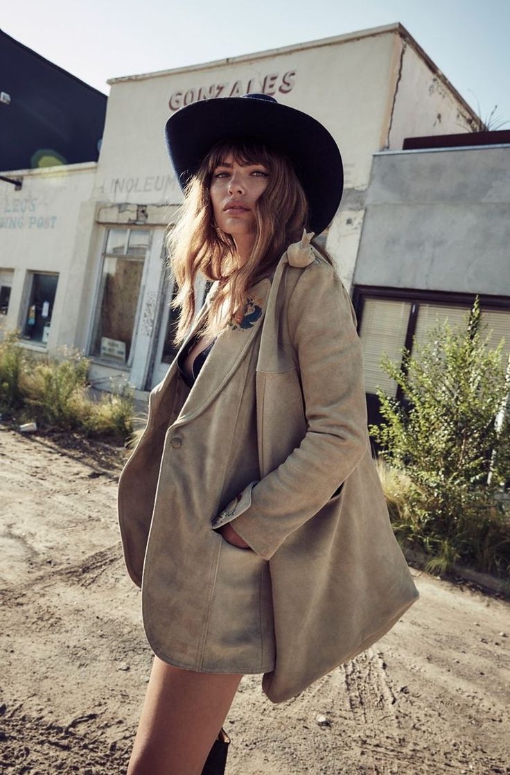 Woman in a beige coat and black hat standing in front of an abandoned building.