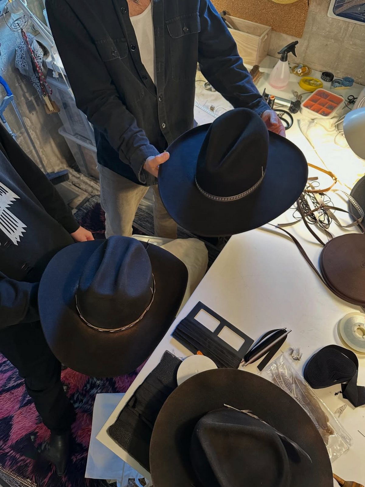 Several wide-brim fedora-style hats in black and dark brown are displayed on a white worktable in a busy craft or workshop setting, with people holding and examining them.