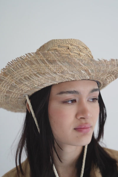 Unisex natural beige straw cowboy hat with a wide frayed, flanged brim, center crease and off-white rope chin strap with blue beads, handcrafted by SoonNoon in Stockholm